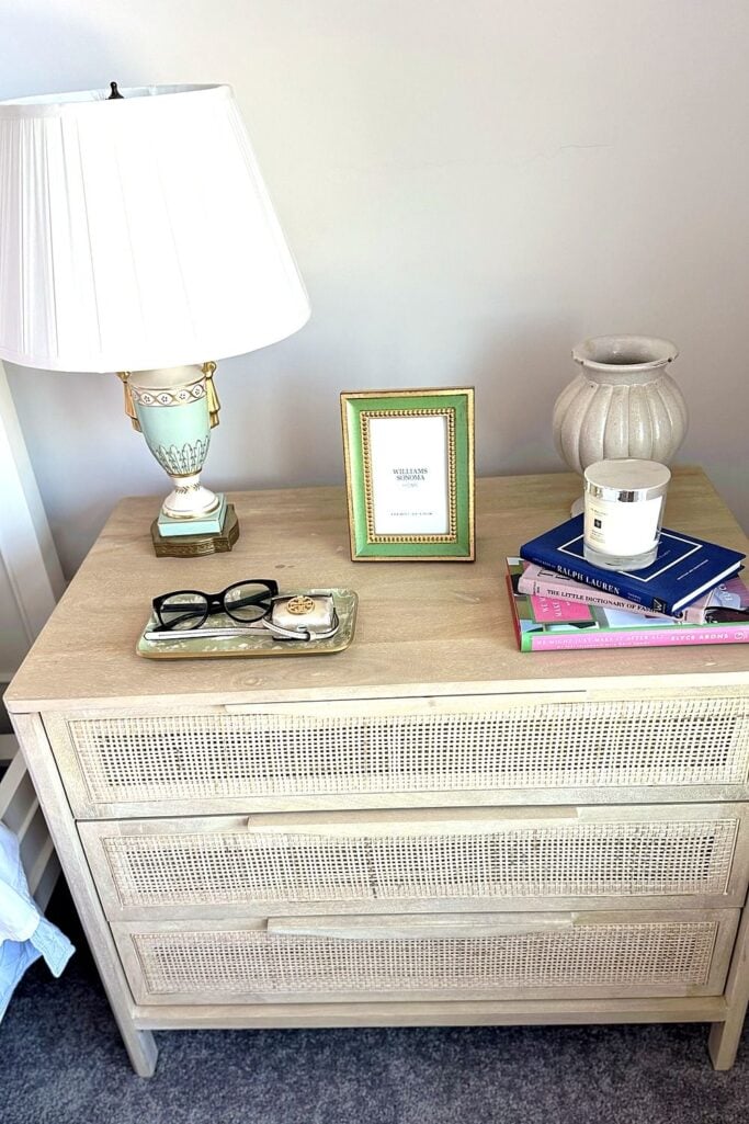 Cane nightstand with a mint urn lamp, green-and-gold frame, candle, and stack of fashion books