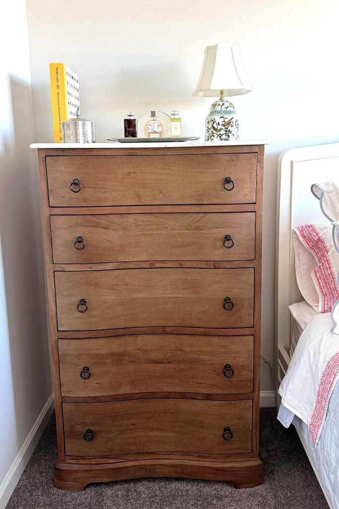 Tall wood dresser with a marble top, perfume tray, and chinoiserie lamp beside the bed